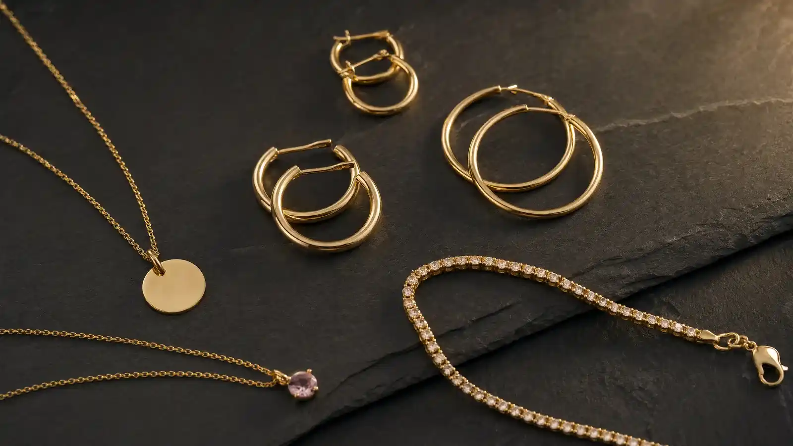 Gold filled jewelry flat lay with hoop earrings, an initial disc pendant, a tennis bracelet and a pink gemstone drop necklace on dark slate
