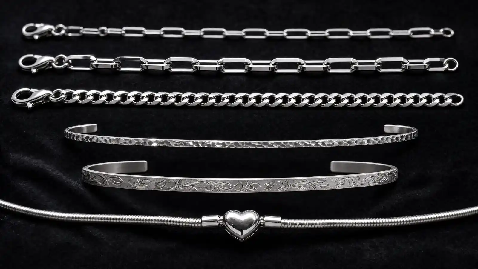 Six sterling silver bracelets laid in parallel rows on black velvet: a fine 3mm paperclip chain, a wider mirror-polished paperclip chain, a curb link chain, a thin hammered open cuff, an engraved floral bangle and a snake chain with a heart-shaped clasp