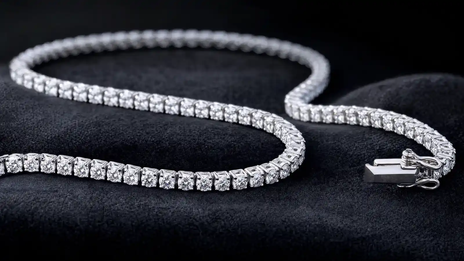 A single white-metal diamond tennis bracelet laid in a gentle S-curve on dark charcoal velvet, showing the continuous line of matched round stones in four-prong settings and the box clasp with its figure-eight safety catch